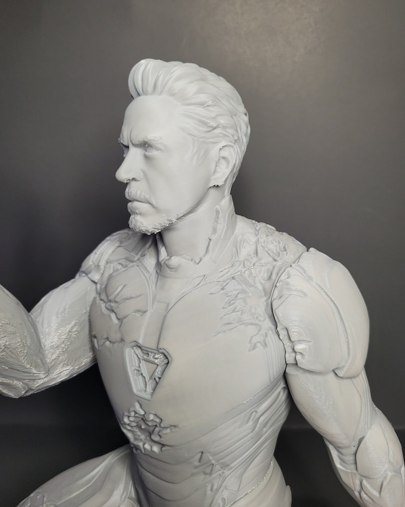 May include: A gray 3D printed figurine of a superhero wearing a detailed, textured suit. The figure has a determined expression and a beard.