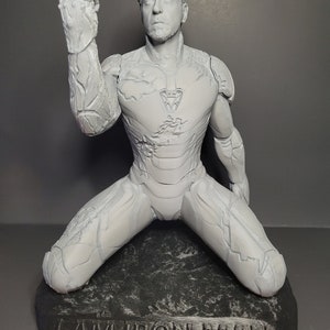 May include: A white 3D printed figurine of Iron Man kneeling on a black rock base. The figurine is in a distressed, weathered style. The base has the words "I AM IRON MAN" engraved on it.