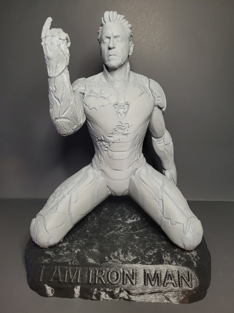 May include: A white 3D printed figurine of Iron Man kneeling on a black rock base. The figurine is in a distressed, weathered style. The base has the words "I AM IRON MAN" engraved on it.