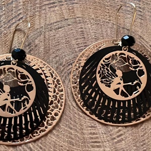 May include: A pair of gold-tone earrings with black enamel and a cutout fairy design. The earrings are round and have a layered look.