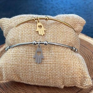 Può includere: Due braccialetti rigidi dorati e argentati con un ciondolo a forma di mano di hamsa. I braccialetti sono esposti su una superficie di tessuto beige.