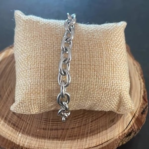 May include: A silver chain bracelet with a curb link design. The bracelet is displayed on a small, beige fabric pillow.