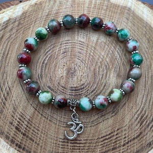 Puede incluir: Un brazalete de jade verde, rojo y blanco con un dije de Om plateado. El brazalete está hecho de cuentas redondas y está enhebrado en un cordón plateado.