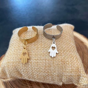 Può includere: Due anelli regolabili con ciondoli a forma di mano di Hamsa. Uno è dorato, l'altro argentato. Ogni ciondolo presenta un cuore ritagliato. Gli anelli sono esposti su un cuscino beige.