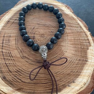 Peut inclure: Un bracelet en perles de lave noire avec un charme argenté représentant un symbole. Le bracelet est doté d'un cordon de fermeture en brun.