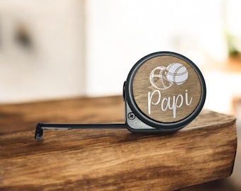 Peut inclure: Un mètre ruban noir et argenté avec une face en bois. La face présente un motif de ballon de basket et le mot "Papi" en écriture blanche. Le mètre ruban repose sur une surface en bois.