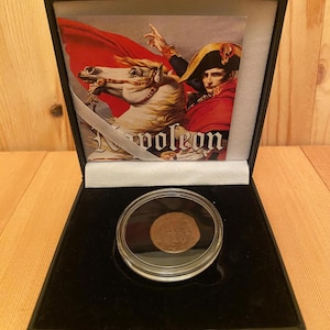 May include: A circular copper coin encased in a clear protective capsule, presented inside a black display box. The box lid features an illustration of Napoleon on a white horse with the text Napoleon printed in white at the bottom of the image.