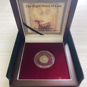 May include: A wooden box with a red velvet interior, containing a silver coin in a clear plastic case. The coin features a hand reaching down from the sky, with the text "The Right Hand of God" and "Medieval German "1 Heller" Silver Coin" on the box.