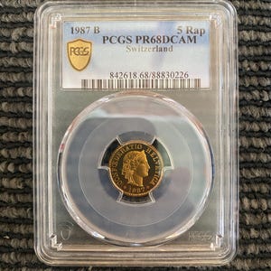 May include: A gold coin in a clear plastic holder with a black label that reads "PCGS PR68DCAM 5 Rap Switzerland 842618.68/88830226". The coin features a woman's profile with the text "CONFOEDERATIO HELVETICA 1887" around the edge.