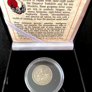 May include: A silver coin from Japan, featuring a chrysanthemum, a phoenix, and a cherry blossom. The coin is in a clear plastic case, sitting on a black surface. The text on the card reads "The Imperial Japanese Silver Coin - 50 Sen".
