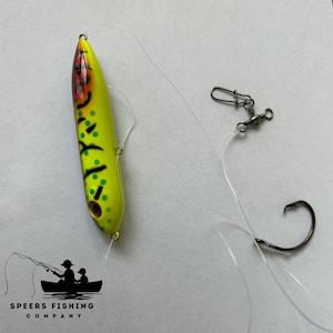 May include: A yellow and green fishing lure with black markings and red accents. The lure is attached to a clear fishing line with a swivel and a black hook.