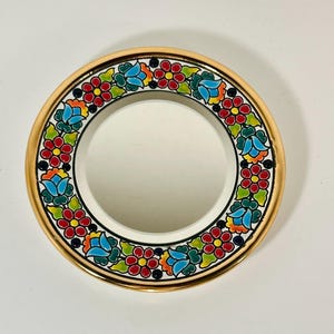 May include: A round, decorative plate with a white center and a gold rim. The rim is adorned with a vibrant floral pattern featuring red, blue, and green flowers. The plate is likely ceramic or porcelain.