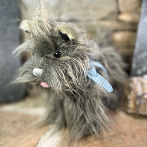 May include: A plush gray terrier dog with a blue and white checkered ribbon around its neck. The dog has a long, shaggy coat, black eyes, and a pink tongue. The toy is made of soft, fluffy material.
