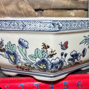 May include: Rectangular ceramic planter with a cream-colored base, adorned with blue floral and butterfly motifs. The rim features a blue geometric pattern. The planter is approximately 8 inches long.