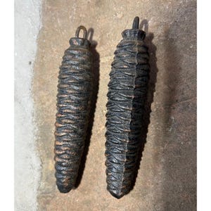 May include: Two antique clock weights, shaped like pine cones, in shades of brown and black. Each weight has a loop at the top for hanging. The weights have a textured, ridged surface.