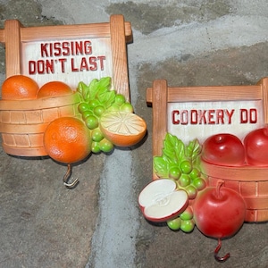 May include: Two decorative kitchen wall hooks. One features a sign that reads "KISSING DON'T LAST" with oranges, grapes, and a lemon. The other hook has a sign that reads "COOKERY DO" with apples and grapes. Both have a hook for hanging.