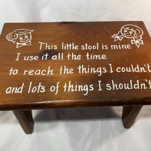 May include: A rectangular brown wooden stool with four legs. The top surface features white painted illustrations of a boy and a girl, along with the text: This little stool is mine, I use it all the time, to reach the things I couldn't, and lots of things I shouldn't.