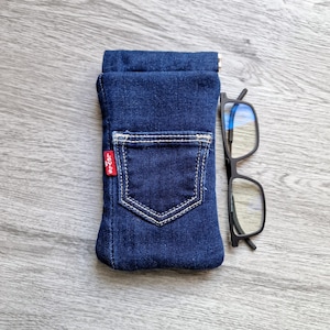 May include: A dark blue denim glasses case with a back pocket design and a red Levi's tag. A pair of black-framed spectacles sits beside the case. The case is made of denim fabric.