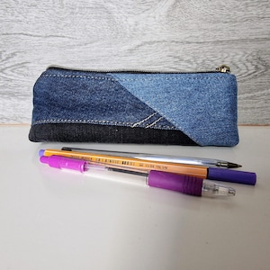 May include: A denim pencil case with a zipper closure, featuring a patchwork design of various shades of blue denim and black denim. Three pens, including a purple pen, are in front of the case.