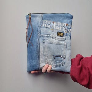 May include: A denim clutch made from recycled jeans. The bag features a zipper closure, a brown leather pull, and a back pocket with decorative rivets. The design incorporates various shades of blue denim.