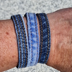 May include: Three denim bracelets in blue shades, stacked on a person's wrist. The bracelets are made from repurposed denim fabric and have visible stitching.