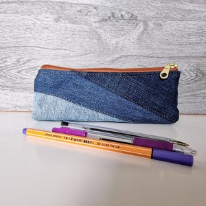 May include: A denim pencil case with a brown zipper and gold pull sits on a white surface. The case is made of different shades of blue denim. Several pens and pencils are in front of the case.