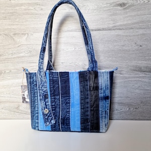 May include: A denim tote bag with vertical stripes in various shades of blue and black. The bag has two handles and a button closure. The bag is made from recycled denim.