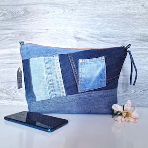 May include: A denim patchwork pouch with a zip closure and a small pocket. The pouch is made from various shades of blue denim and has a brown zip. A smartphone and a flower are in the foreground.