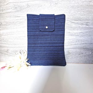 Könnte beinhalten: Eine rechteckige blaue Stoffhülle mit einem Klappenverschluss, der mit einem silbernen Druckknopf gesichert ist. Die Hülle hat ein strukturiertes Streifenmuster. Der Hintergrund zeigt eine weiße Oberfläche und eine graue Holzwand.