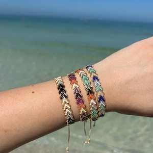 May include: Five colorful chevron patterned friendship bracelets with gold beads on a person's wrist. The bracelets are in shades of white, black, orange, purple, and blue.