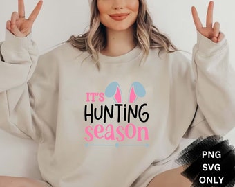 May include: A light beige sweatshirt with the text "It's Hunting Season" in pink and black lettering. The text is accompanied by a pair of blue bunny ears with pink insides.
