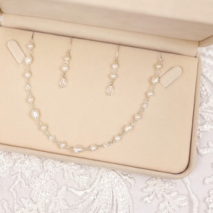 Peut inclure: Un collier et des boucles d'oreilles en perles dans un coffret à bijoux beige. Le collier est composé de petites perles blanches et est présenté sur un fond en velours beige. Les boucles d'oreilles sont également composées de petites perles blanches et sont présentées sur un fond en velours beige.