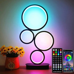 Pode incluir: Um candeeiro de mesa moderno com quatro luzes LED circulares que mudam de cor. As luzes são dispostas em cascata, com a maior na parte superior e a menor na parte inferior. O candeeiro tem uma base preta e um comando remoto.