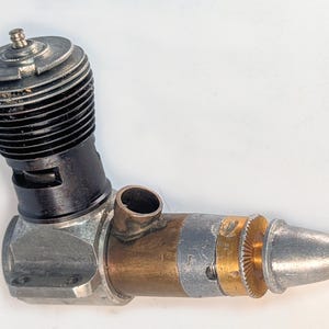 Może przedstawiać: Mały silnik spalinowy do modeli pojazdów z czarną żebrowaną głowicą cylindra, srebrną metalową skrzynią korbową oraz mosiężnym zespołem wlotowym ze srebrnym stożkiem nosowym. Silnik charakteryzuje się kompaktową konstrukcją mechaniczną.