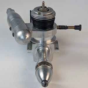 May include: A silver-coloured internal combustion engine for a remote-controlled vehicle, featuring a metallic cylinder head with black cooling fins, a side-mounted exhaust muffler, a brass-coloured needle valve assembly, and a front-facing propeller hub.