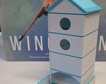 May include: A white and light blue dice tower with a textured wood grain finish. The tower features a peaked roof, two circular openings on the front, and a rectangular base tray with a light blue floor for catching dice during board games.