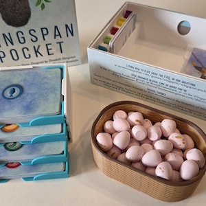 May include: The Wingspan Pocket board game set includes a box with the title, a white card organizer tray holding stacks of cards with blue, orange, green, and red icons, a white storage box with game instructions, and a brown oval bird egg bowl.