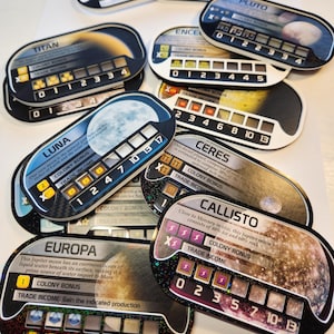 Pode incluir: Uma coleção de cartas de jogo coloridas com corpos celestes como Titã, Luna e Europa. Cada carta tem um design único com texto, números e ilustrações, incluindo indicadores de renda comercial e bônus de colônia. Cartas de jogo.