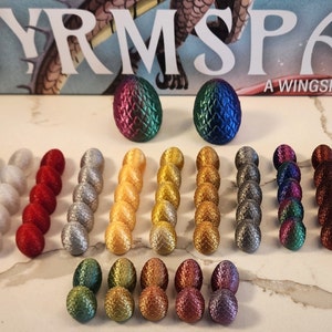 Op de afbeelding: Een set van 24 iriserende draken eieren voor het bordspel Wyrmspan, een Wingspan-spel. De eieren zijn in verschillende kleuren, waaronder zwart, blauw, wit, rood, zilver, goud, groen en paars.