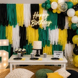 May include: A birthday party setup with a white table, cake, cupcakes, and balloons. The backdrop features black, green, yellow, and white streamers. A neon sign reads "Happy Birthday." A large illuminated number "5" is on the left.