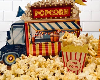 Puede incluir: Un camión de palomitas de maíz rojo, blanco y azul con la palabra "POPCORN" en la parte delantera. Un pequeño contenedor de palomitas de maíz a rayas rojas y blancas con las palabras "POP CORN" está sentado en un montón de palomitas de maíz.