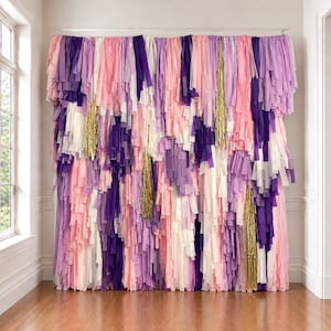 May include: A decorative backdrop made of long, vertical strips of paper in shades of purple, light pink, white, and lavender, interspersed with shimmering gold metallic fringe accents hanging from a horizontal rod across the top.