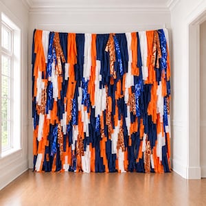 May include: A decorative backdrop made of long, vertical fabric strips in alternating colors of navy blue, bright orange, and white, interspersed with shimmering metallic copper and reflective blue foil ribbons hanging from a top rod.
