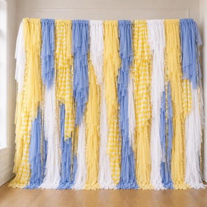 May include: A decorative backdrop curtain featuring vertical layers of tulle and fabric strips in alternating colors of light blue, white, and yellow, including sections with a yellow and white gingham check pattern and metallic gold fringe accents.