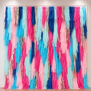 May include: A decorative fringe backdrop featuring vertical strips of fabric in shades of bright pink, light pink, turquoise, and dark blue, interspersed with shimmering gold sequin strands hanging in a layered, multi-colored pattern.