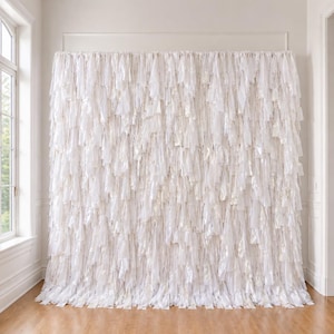 May include: A large, rectangular backdrop composed of numerous vertical strips of white fabric. The material has a slightly translucent and reflective quality, creating a layered, textured appearance that hangs from a horizontal rod.