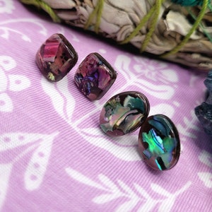 May include: Four resin earrings. Two are square-shaped with embedded pink and purple elements. The other two are oval-shaped with iridescent abalone shell. The earrings are on a lavender fabric with a white floral pattern.