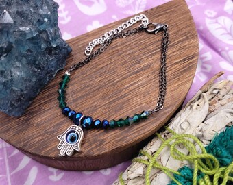 May include: A silver-toned bracelet featuring a Hamsa hand charm with a blue eye design. The bracelet has a dark chain and is adorned with faceted green and blue beads. The bracelet is displayed on a wooden surface.