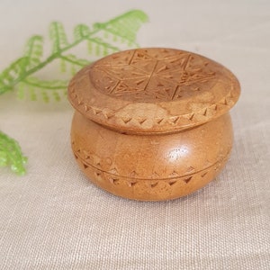 Könnte beinhalten: Kleine, runde Holzbox mit Deckel. Deckel und Boden sind hellbraun und weisen geschnitzte geometrische Muster auf. Die Box dient wahrscheinlich zur Aufbewahrung kleiner Gegenstände. Der Hintergrund ist neutral.