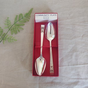 Puede incluir: Dos cucharas de plata con mangos adornados en una caja roja con el texto "COMMUNITY PLATE" en la parte superior.